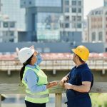 Engineers at a construction site reviewing architectural plans with modern cityscape background.